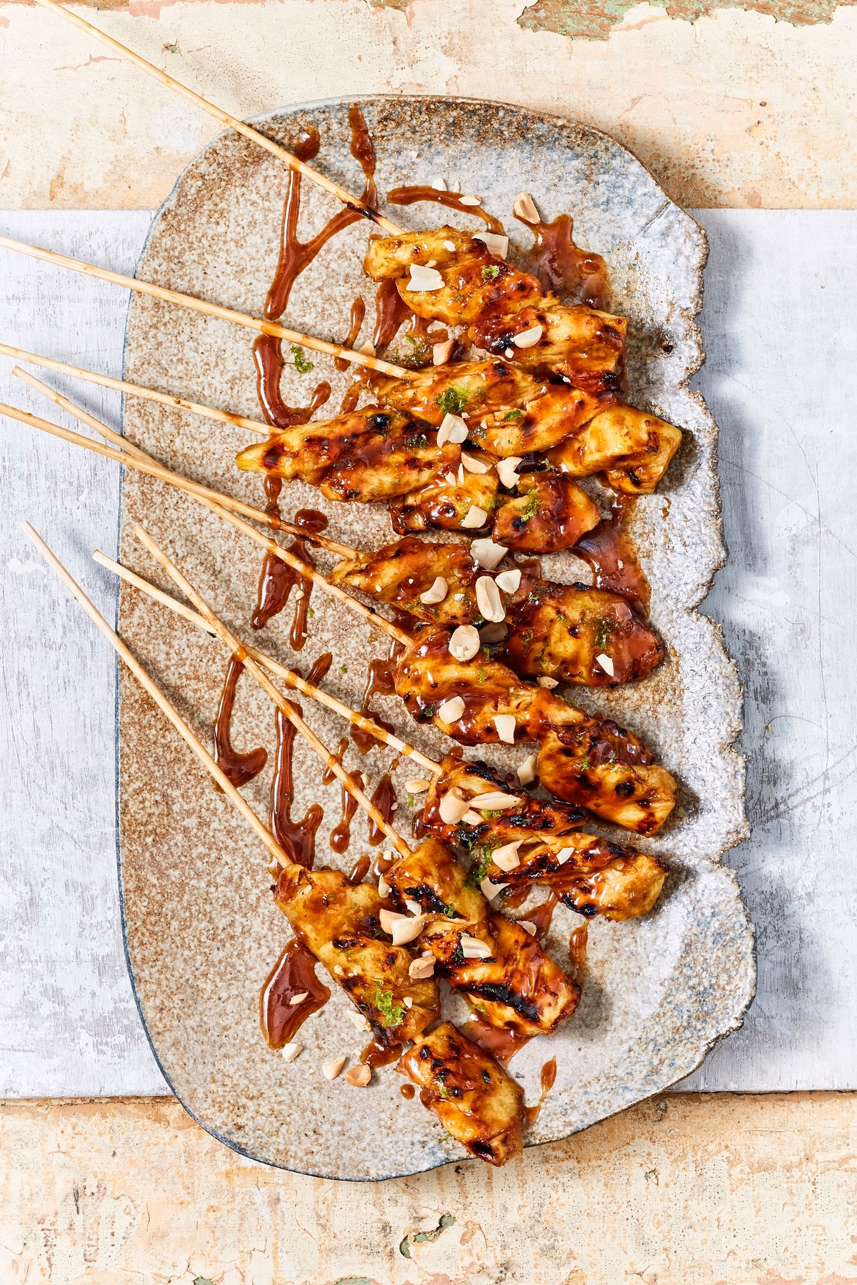 Grilled chicken skewers with sauce and chopped nuts on a textured oval plate, set on a rustic surface.